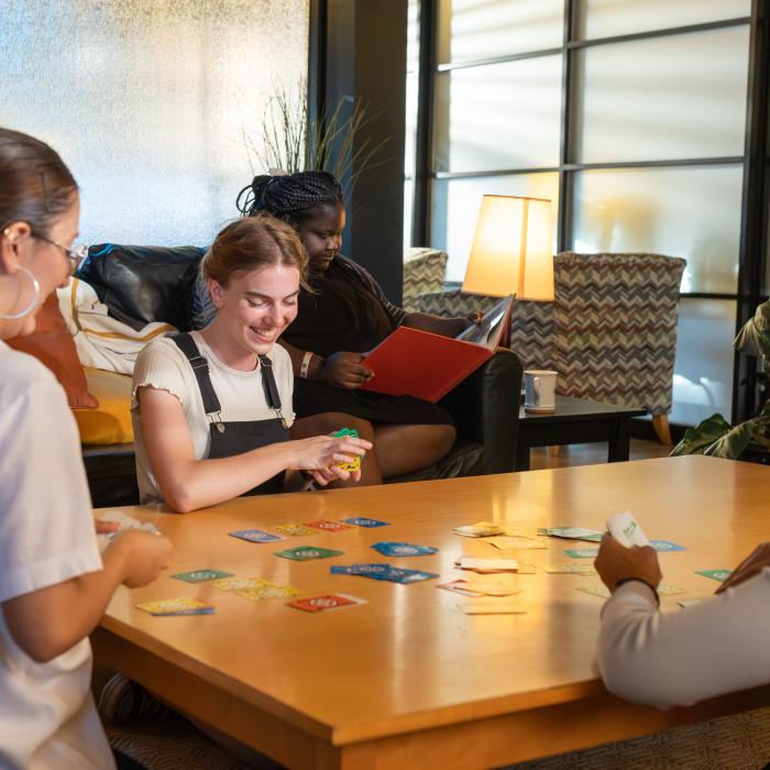 Three women play a card game at a table, one reads on a couch nearby.