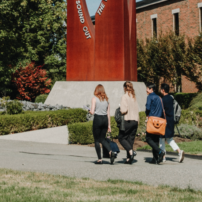 Three people walking on campus path near a brick building and red sculpture.