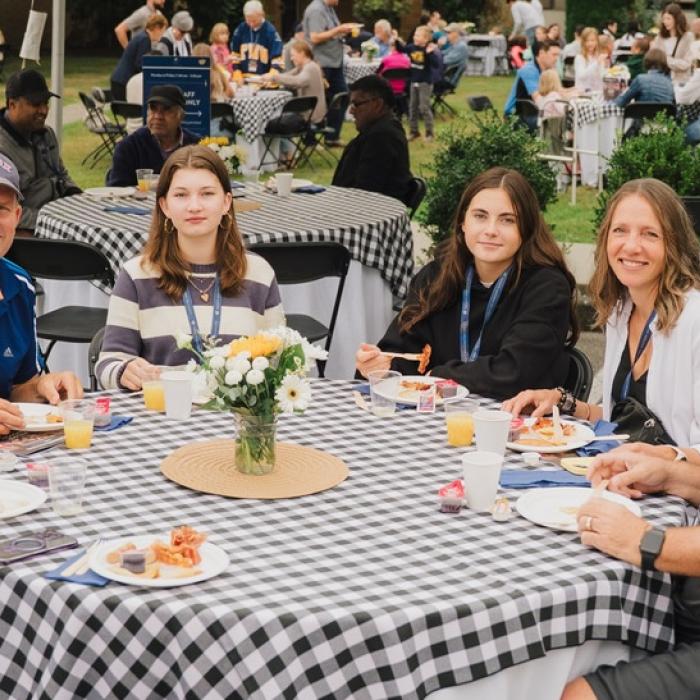 Group of people sitting at an outdoor table with checkered tablecloths, smiling.