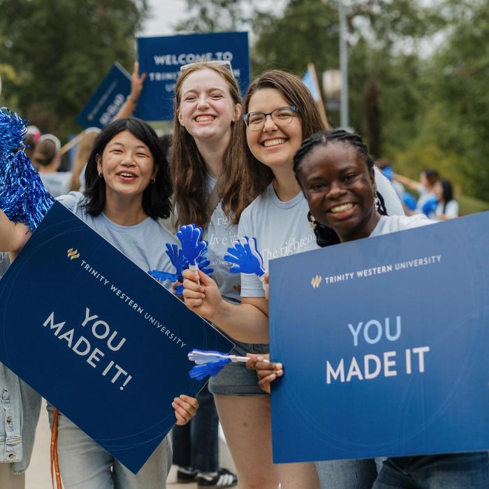 Four people smiling, holding blue signs saying "YOU MADE IT" with balloons nearby.