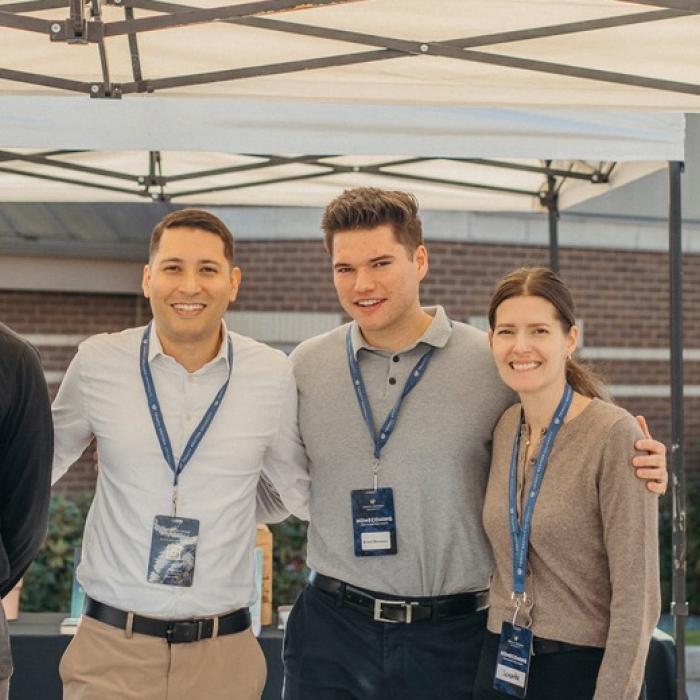 Four people smiling under a canopy with lanyards, next to a sign.