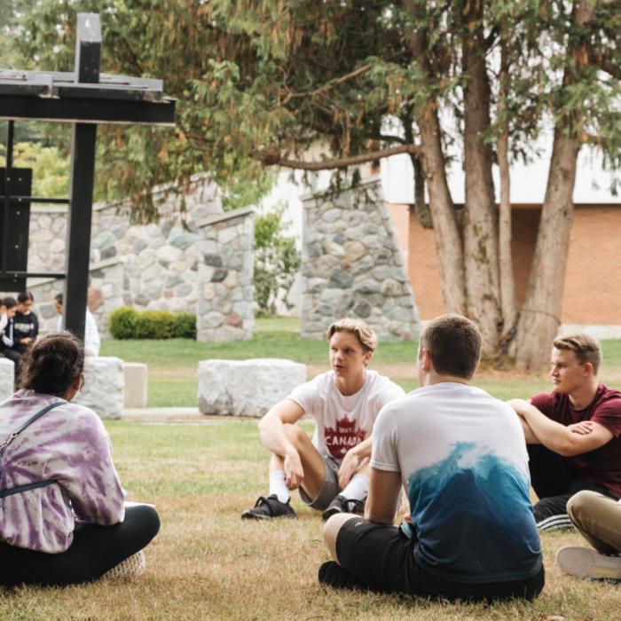 Students sitting in a circle on grass, school building in background.