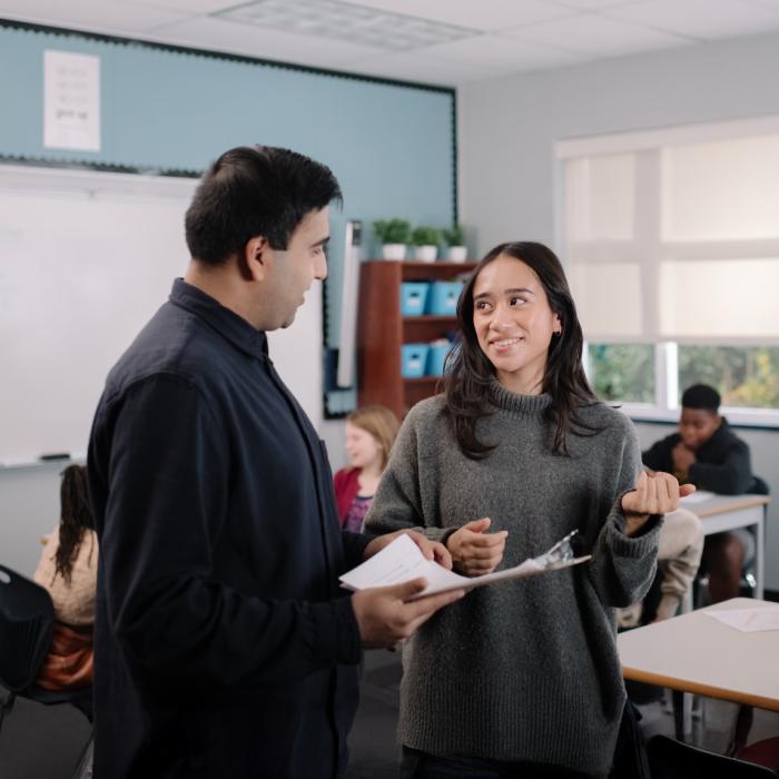 Teacher and student talking in a classroom, others working in the background.