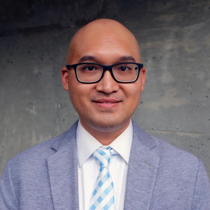 Bald man in glasses wearing a gray suit and blue tie, standing against a concrete wall.