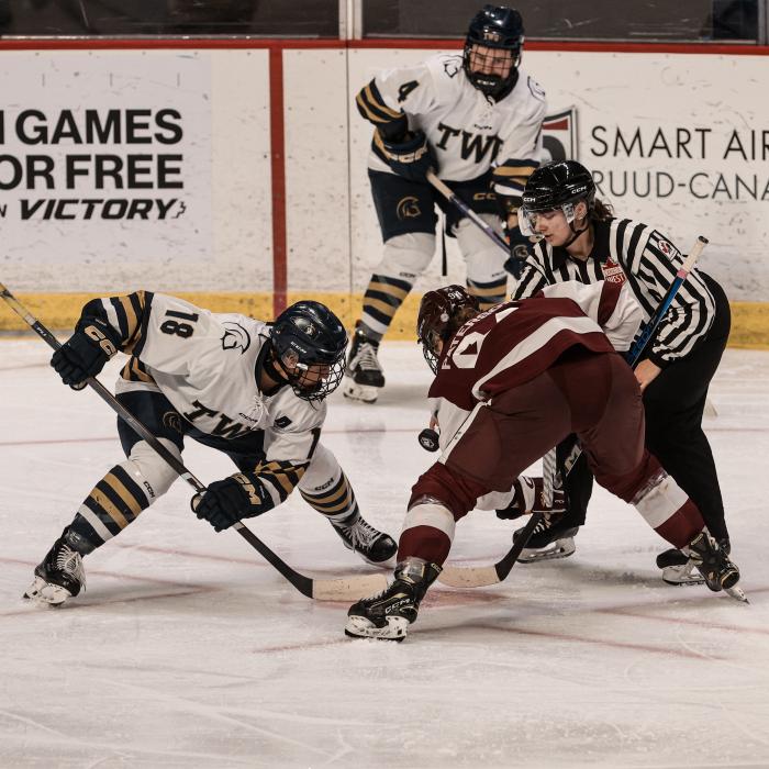 Two hockey players face off on the ice, one in a white TWU jersey and the other in a maroon uniform, with a referee dropping the puck and another player in white positioned behind them. Advertising boards are visible in the background.