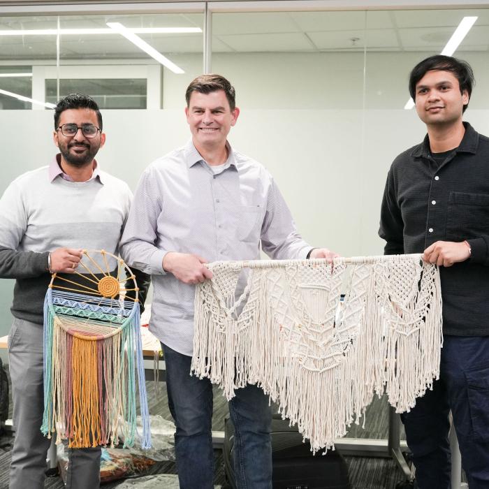Three people stand indoors holding handmade woven artworks, including a colourful dreamcatcher-style piece and a large macramé wall hanging. 