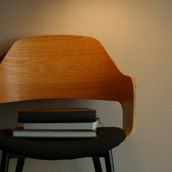A wooden chair with a black cushioned seat sits against a neutral wall, illuminated by soft lighting. A small stack of books rests neatly on the seat.