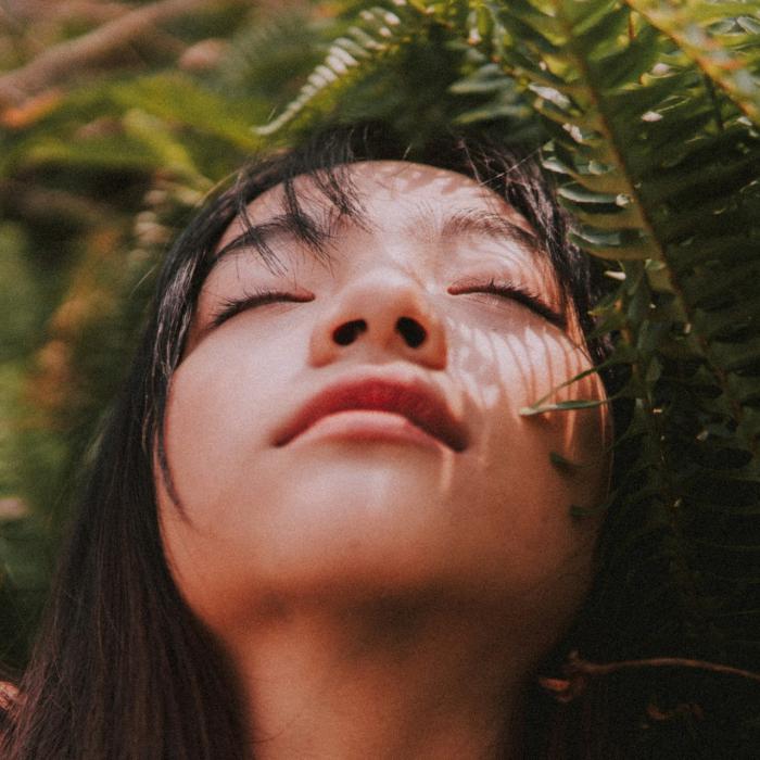 A woman with long dark hair is positioned outdoors among dense, green fern leaves. 