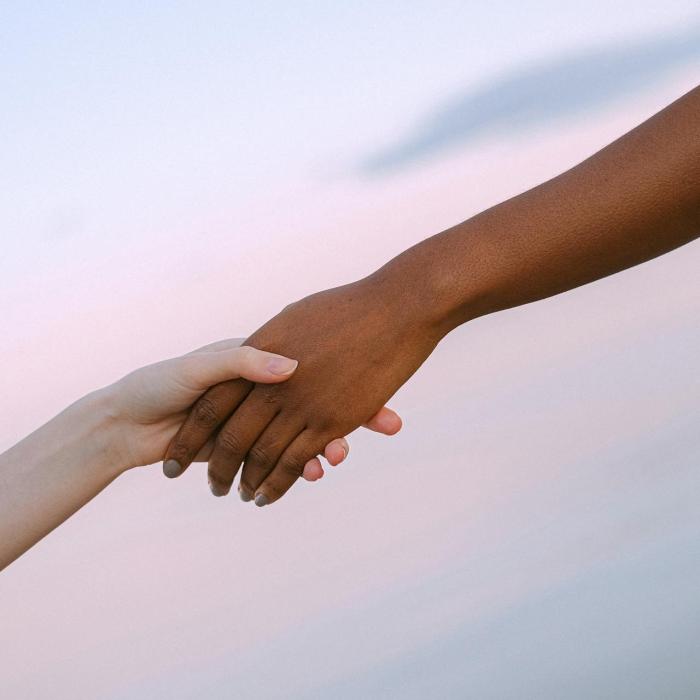 Two hands reaching toward each other and holding against a soft, pastel sky background.