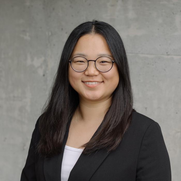 Headshot of Sarah Lee Charlton wearing glasses and a black blazer, standing in front of a grey concrete wall.