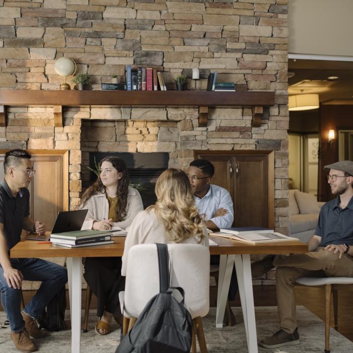 TEDS students gather around a table in the Graduate Collegium, participating in a discussion with laptops, books, and notebooks in a lounge-style learning space featuring a large stone fireplace.