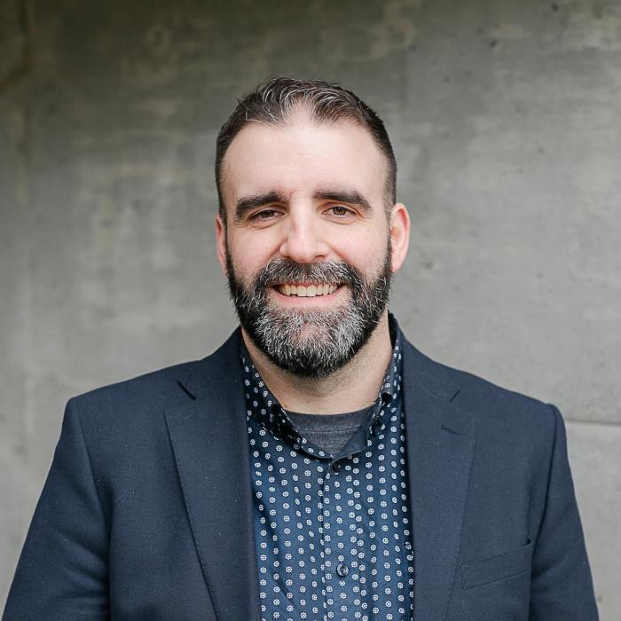 Headshot of Julian Erb wearing a dark blazer and patterned shirt, standing in front of a grey concrete wall.