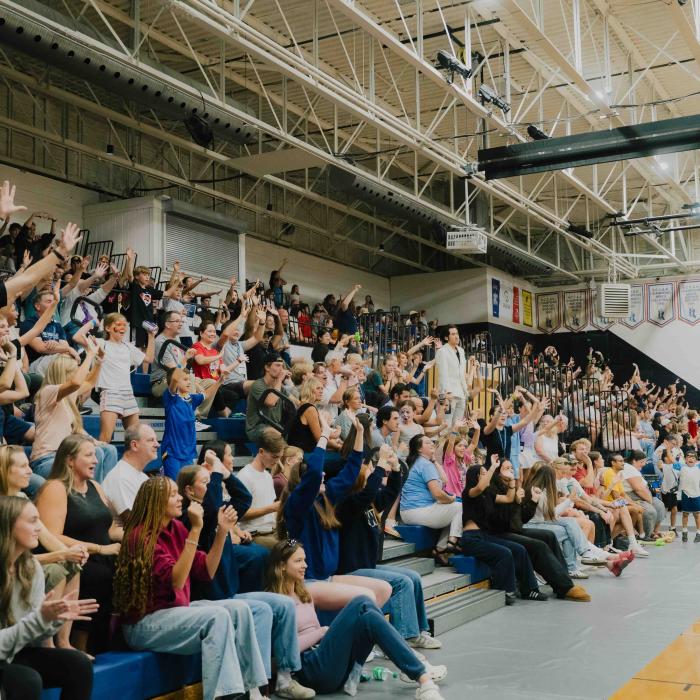 A crowd of adults and children cheer with raised hands from gymnasium bleachers during TWU Homecoming.