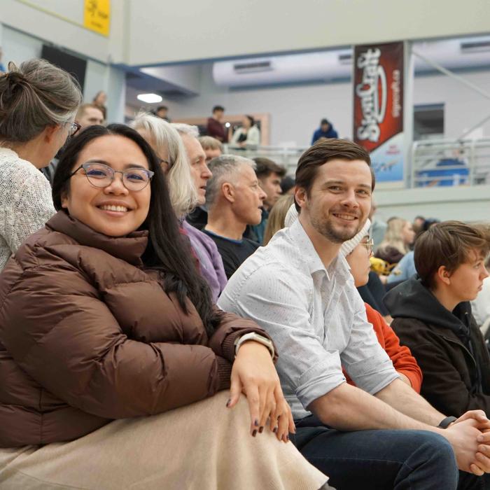 Two attendees smiling at the camera while seated among spectators in gymnasium.