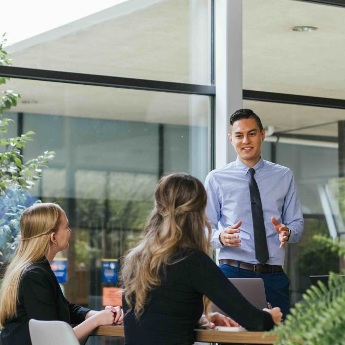 A man standing beside a table speaks with seated colleagues in a bright meeting space with large windows and plants.