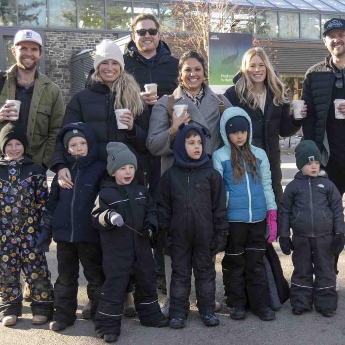 A group of adults and children dressed for cool weather gather outdoors in front of a campus building while holding warm drinks.